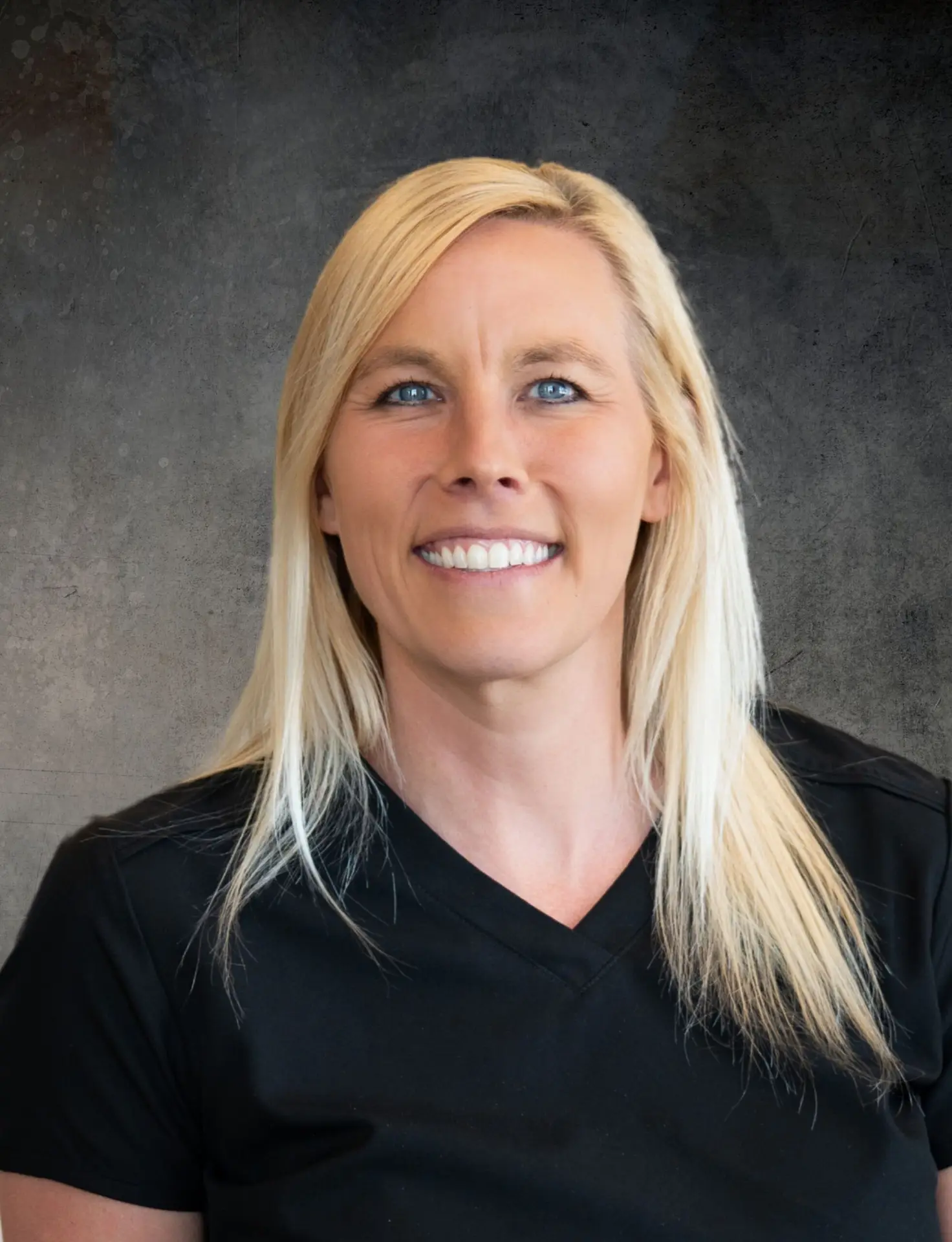 Kim Dewing, PA-C, smiling and wearing black medical scrubs, seated in a warmly lit clinical setting.