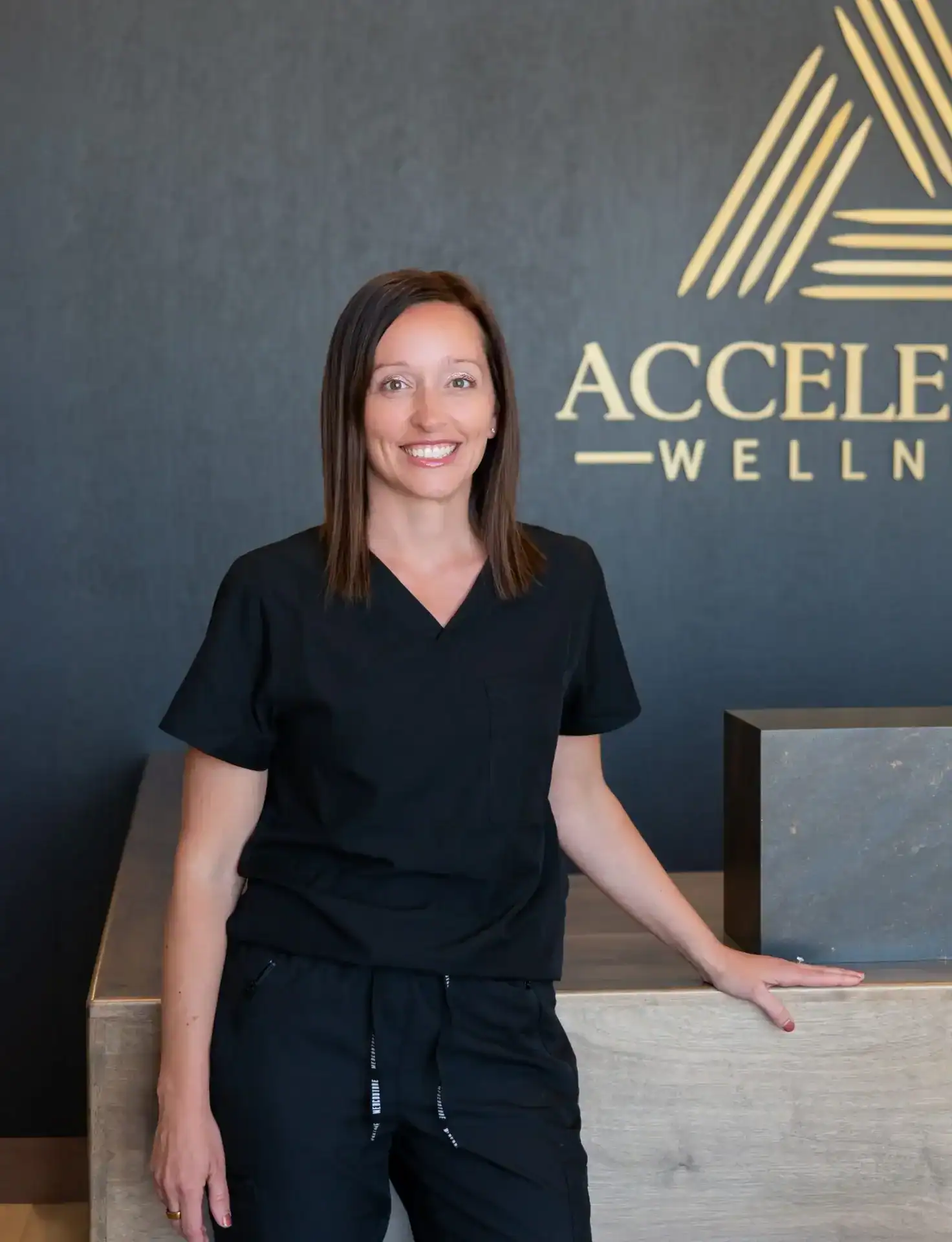 Angela Liddell, CRNA, ABAAHP, smiling in black scrubs in front of the Accelerate Wellness logo wall.